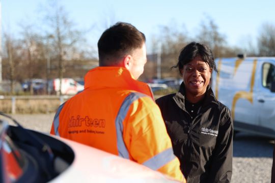 thirteen repairs operative with orange coat on stands with back to camera talking to a colleague who is looking straight into the camera