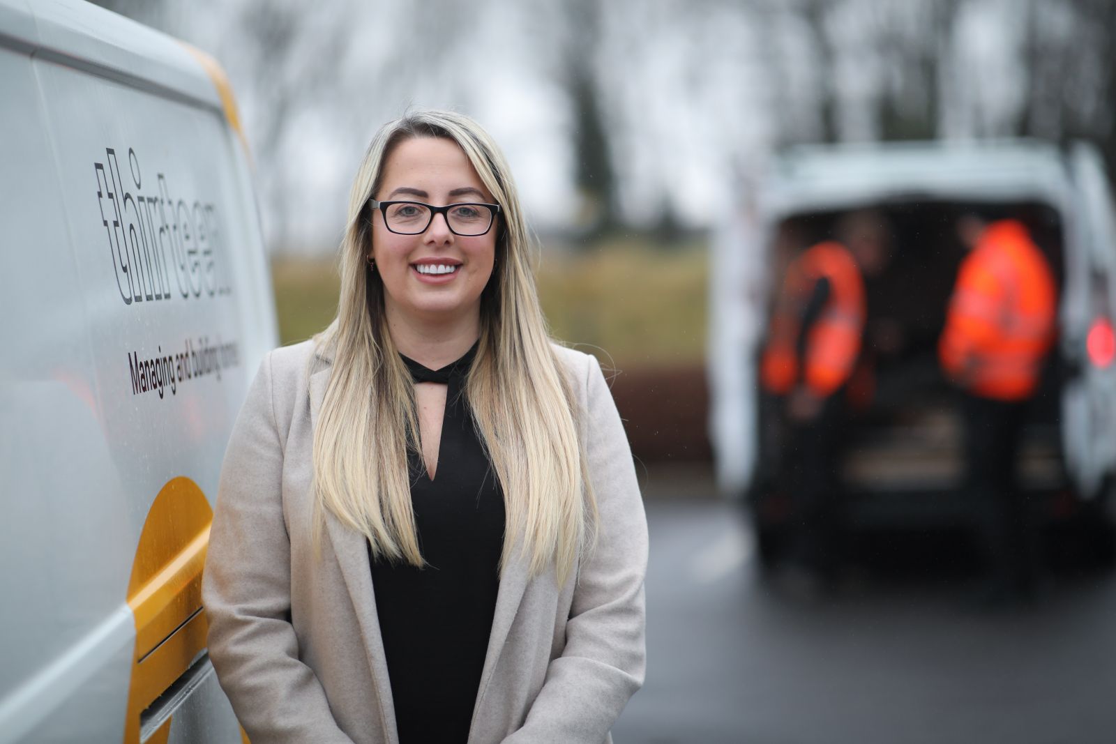 women with long blonde hair wearing black glasses looking into camera standing to the side of a white van with Thirteen logo.