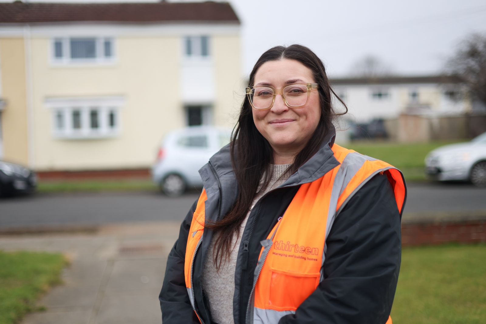 site supervisor in high vis orange jacket, female with long dark hair and glasses looking at the camera standing in front of a cream fronted house