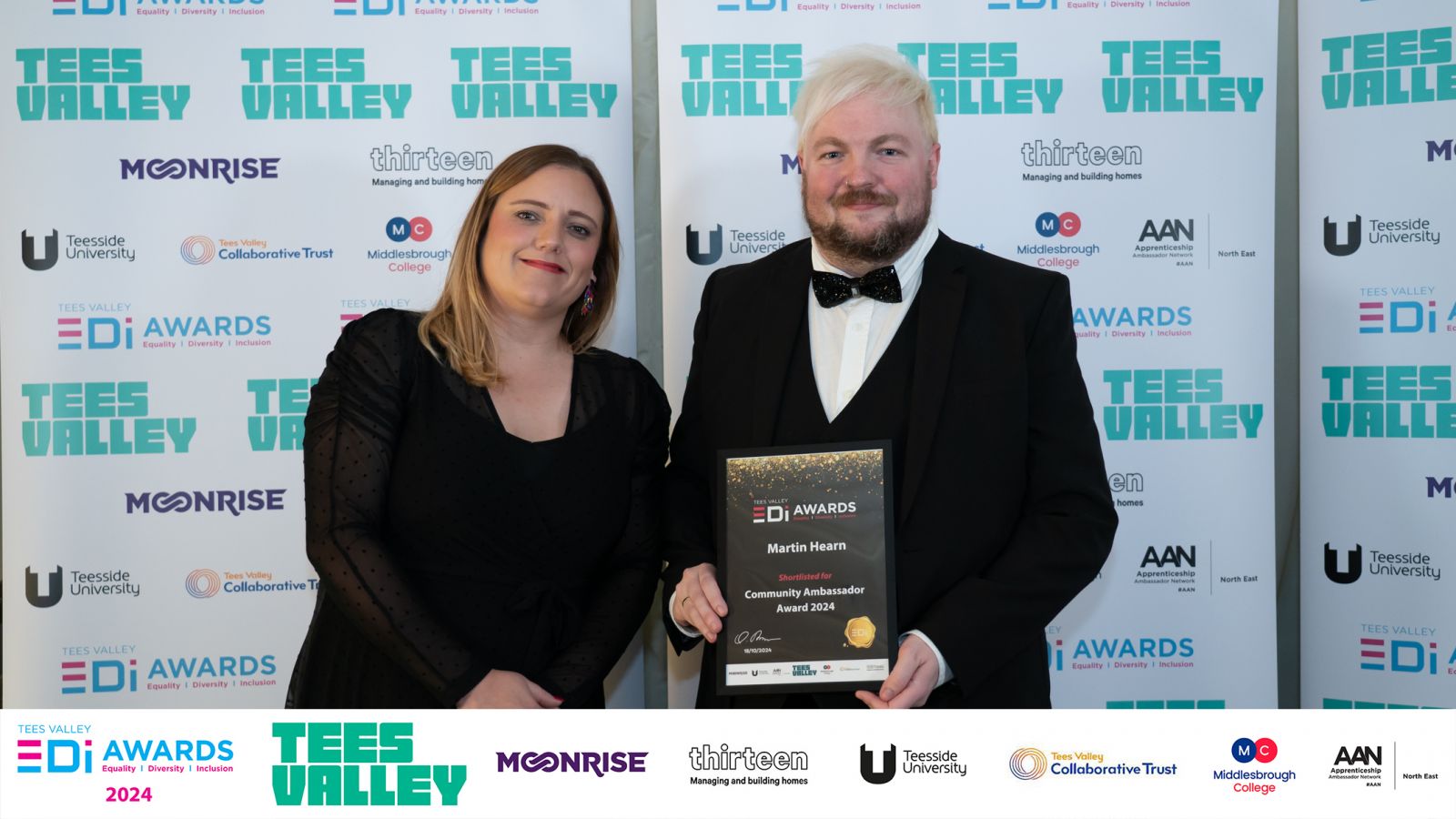 martin hearn standing in front of tees valley advertising boards with his shortlsting certificate for community ambassador. to the left of Martin is a lady with long blonde hair wearing a full length black dress