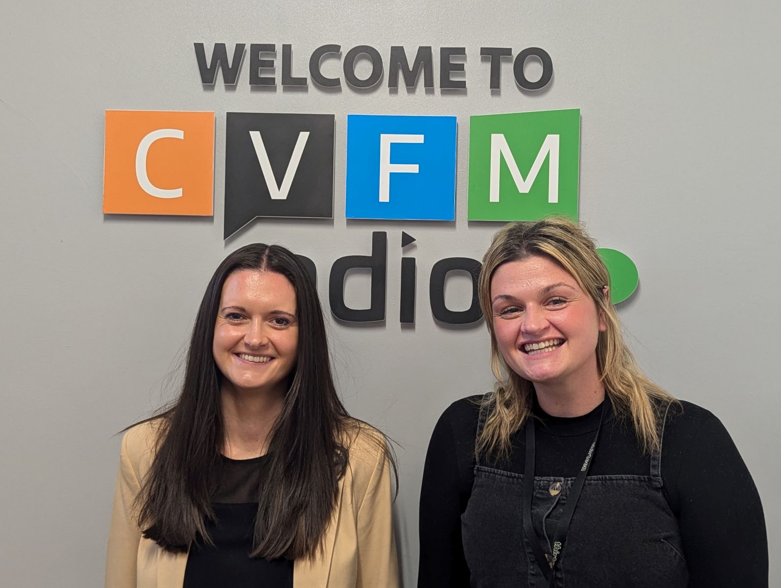 two women one with long dark hair and one with mid-length blonde hair smiling at the camera in front of a grey walll with welcome to CVFM radio on it