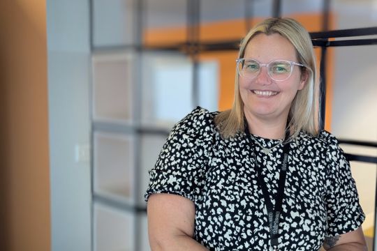 female colleague at hudson quay standing in front of room dividers. Female wearing clear rim glasses, has blonde hair and is wearing a black and white dress