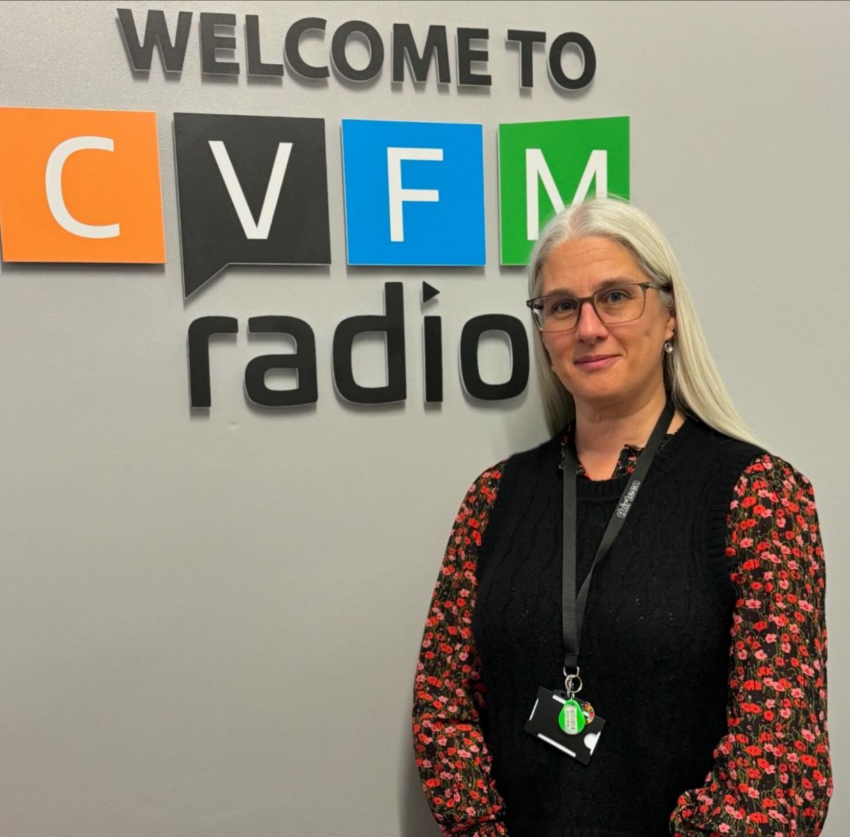 Sara Wetherill, blonde hair and glasses wearing a black sleeveless jumper with a red floral sleeved blouse stood in front of a grey wall with welcome to cvfm radio on it.