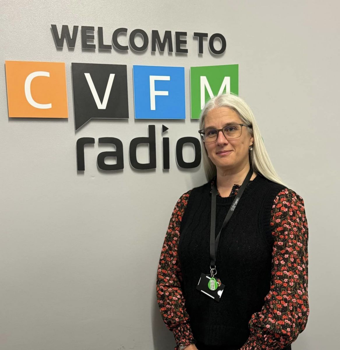 Sara Wetherill, blonde hair and glasses wearing a black sleeveless jumper with a red floral sleeved blouse stood in front of a grey wall with welcome to cvfm radio on it.